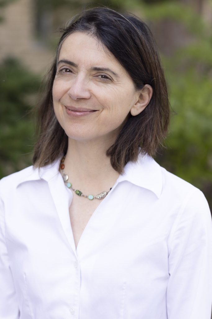 Photo of Alex Huneeus, a light skinned woman with brown hair. She wears a white shirt and smiles at the camera.