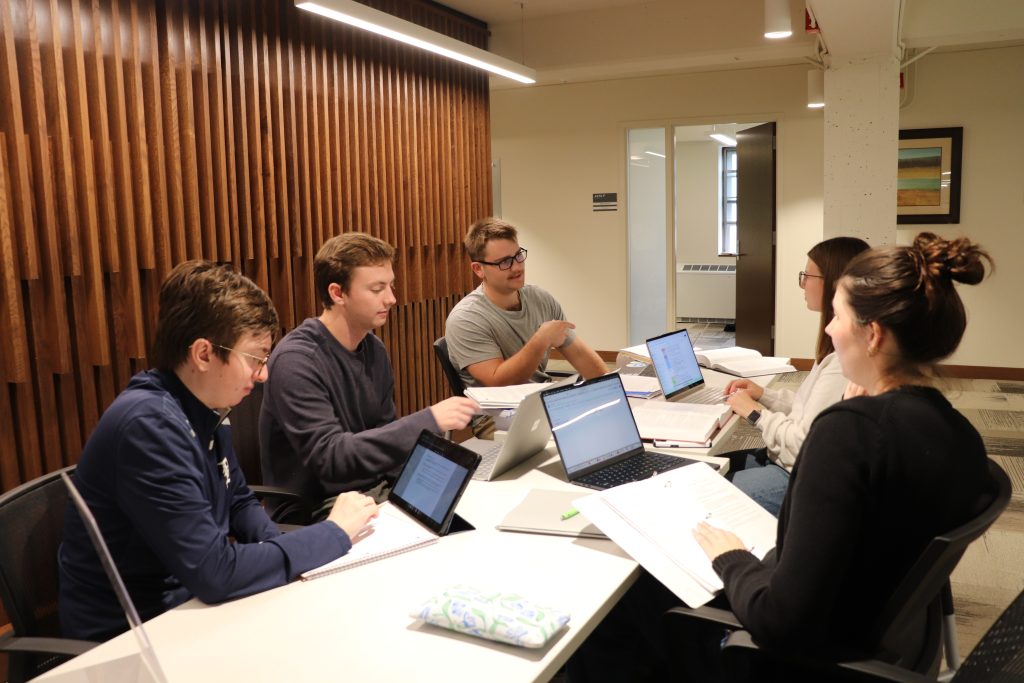 Students seated at a long conference table with a decorative wood wall on one side.