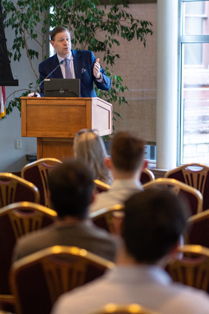 A middle aged man with fair skin wearing a blue suit and gray tie gestures to the crowd as he speaks.