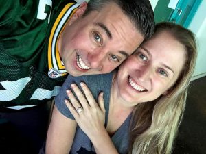 Photo of Ryan Packer in his Packers jersey smiling alongside his then-fiancé Marie Packer as she smiles and shows off her engagement ring.