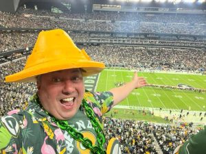 Ryan Packer gesturing to the football game while wearing his Packers memorabilia and a cheese hat.