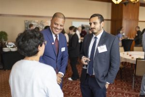 A black man in a suit, and a brown skinned man in a suit, speak with a woman.