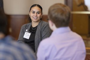 A woman with brown skin and dark hair and wearing a warm cardigan speaks to someone in the foreground.