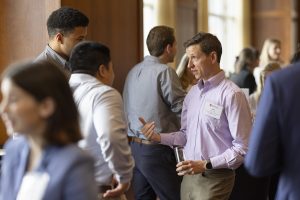 A man in a light plum dress shirt holds a glass and talks with two men nearby, all in a crowd.