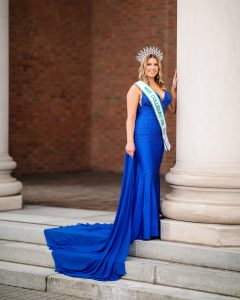 A fair skinned blonde woman in an elaborate blue ball gown and decorative crown stands beside a stone column.