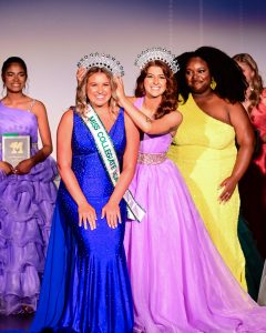 A diverse group of women stand together while wearing colorful, elaborate gowns. One woman in a crown is placing a crown on another woman. They are all smiling.