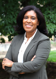 Photo of Emma Shakeshaft, a middle-aged woman with a glowing bronze complexion, black hair and dark eyes; she smiles to the camera and is wearing a gray suit.