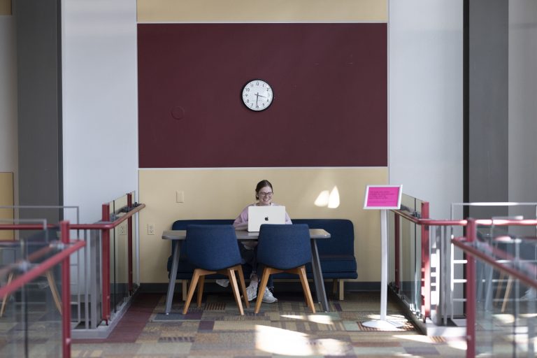 Young woman sits at a table with her laptop in the upstairs of the Law School Atrium.