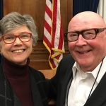 Two senior folks standing together - a woman with gray hair and red glasses and a bald man with brown glasses
