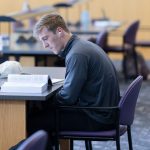 A young blonde man sits with earbuds in his ears while reading from a large book