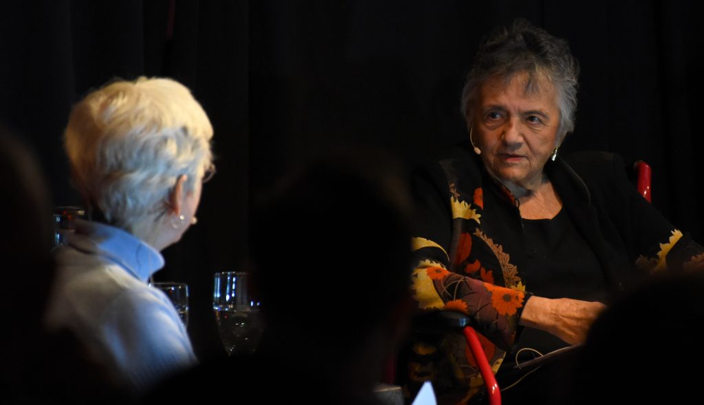 To senior women speak on a dark stage.