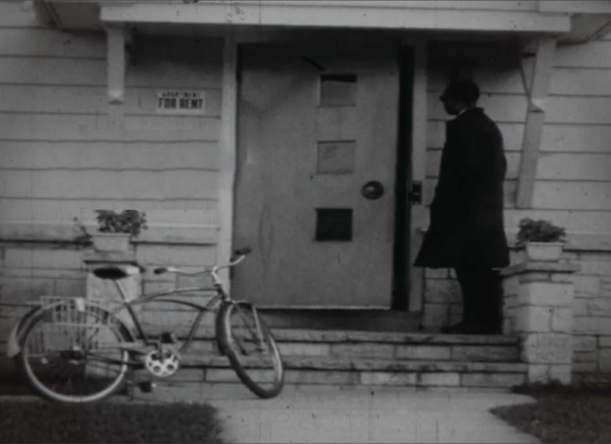 black and white photo of a man in front of a house with a bike outside