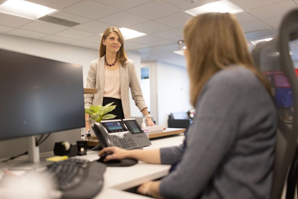 A woman is speaking to another who is seated behind a desk.