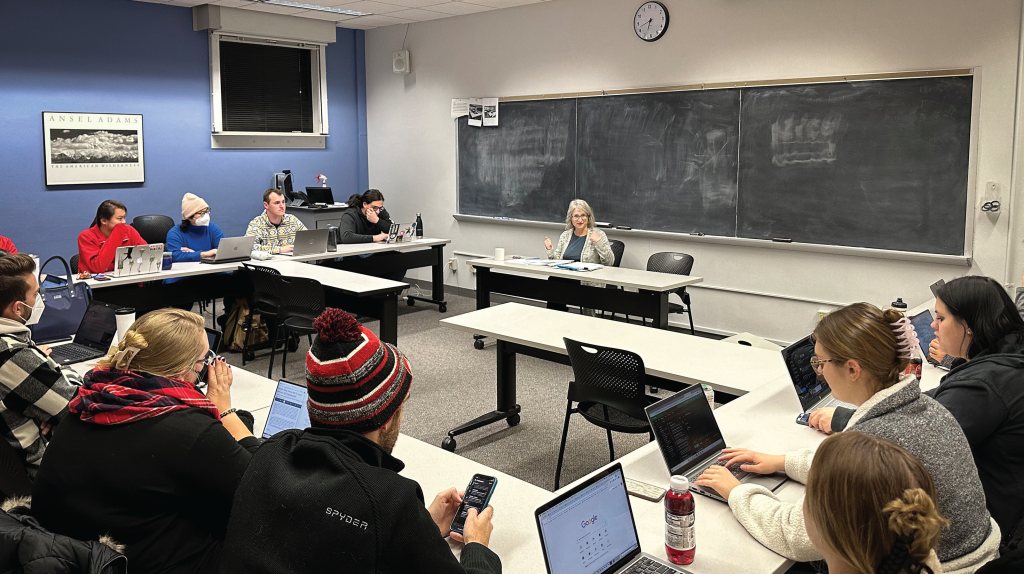 Professor Margaret Raymond speaks to a class while sitting in front of a chalkboard, several of the students are wearing medical masks as they type on laptops and phones.