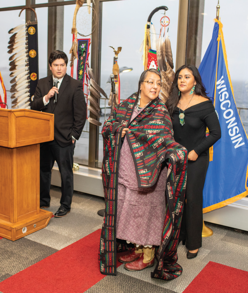 Three people in the image. One person speaking at the podium with a microphone. One person is standing behind a third draped with a blanket.
