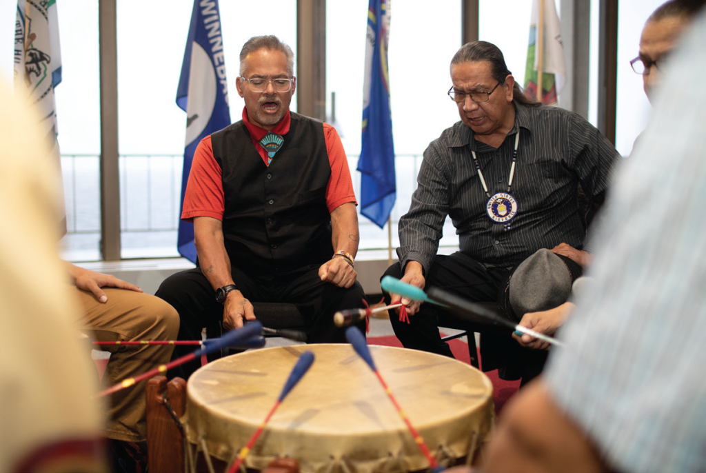 Several people hitting a drum with colorful sticks while singing.