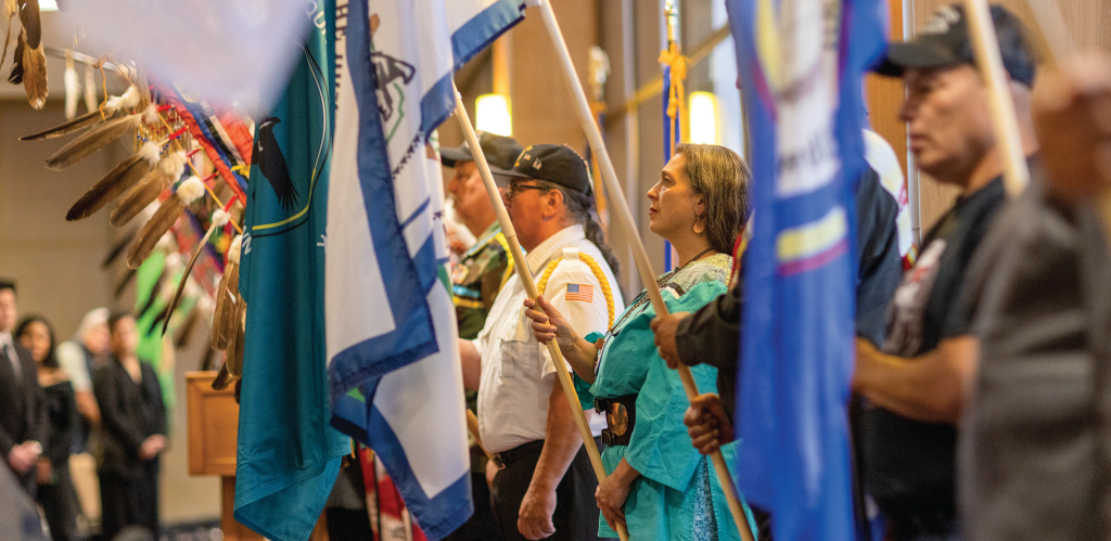 Several people in uniforms and traditional clothing standing in a line while holding flags with feathers attached.