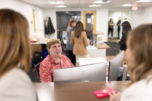 Braden Kundert, a second-year student at the University of Wisconsin Law School, works as a student ambassador for the Admissions team. He works closely with staff, other ambassadors and prospective students. In the image, he is seated behind a computer at a service desk and is speaking with coworkers while smiling.