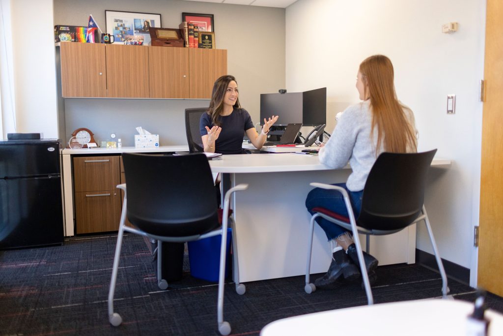 A woman in black sits at a desk while gesturing with her hands as she talks to another woman in a chair.