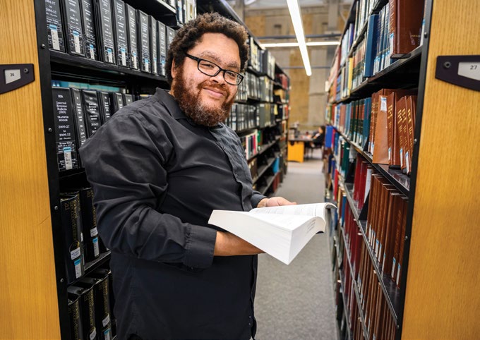Photo of Steven Wright standing between law library book stacks while holding a book. Photo taken by Bryce Richter.