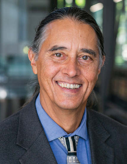 A headshot photo of Heinz Klug, wearing a dark gray suit, blue shirt, and striped tie, while smiling at the camera.