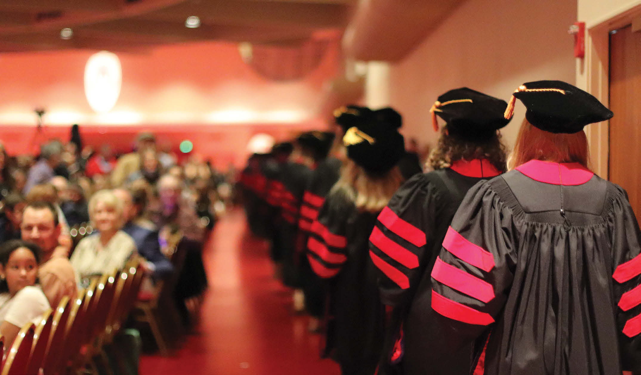 A line of graduating law students are lined up, while wearing their black caps and robes with red arm stripes, behind a seated crowed who watches them with anticipation.