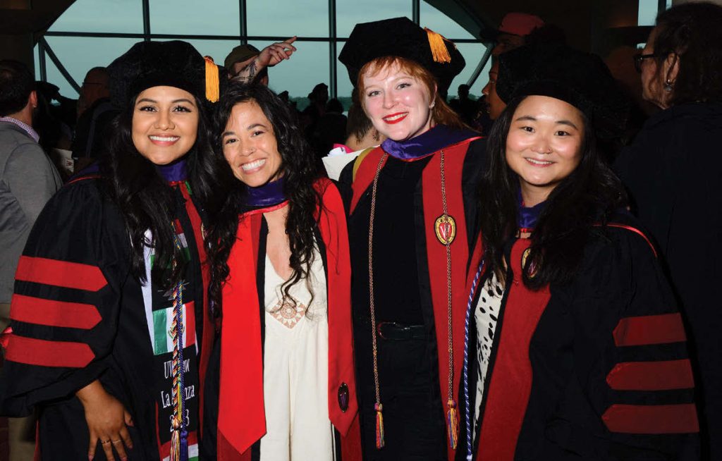 A diverse group of female graduates smile at the camera while wearing variations of UW Law graduation robes and caps.