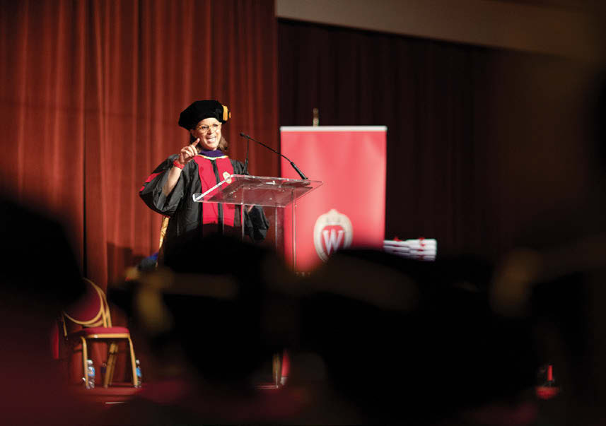 Erin McBride, a woman in graduation garb, stands at a podium and addresses the crowd at commencement.