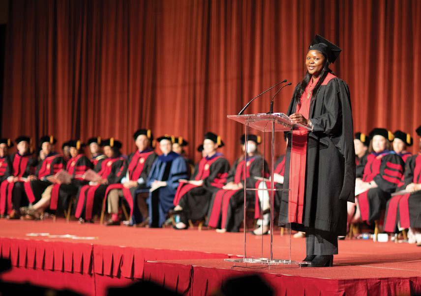 Harriet Maona, a black female graduate, speaks from a podium in front of other graduates while wearing graduation robes and cap.