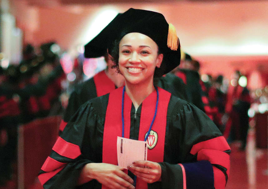 A graduating law student of color smiles at the camera as she holds a slip of paper while wearing black and red robes and a black cap.