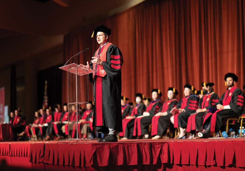 A man in graduation garb, Trager Metge, stands at a podium in front of other graduates on a stage and speaks to the audience.