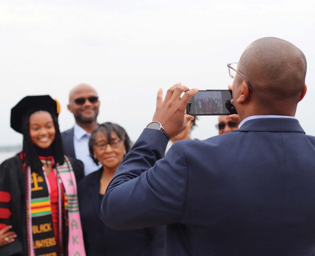 A woman of color wearing graduation robes takes a photo with her family.