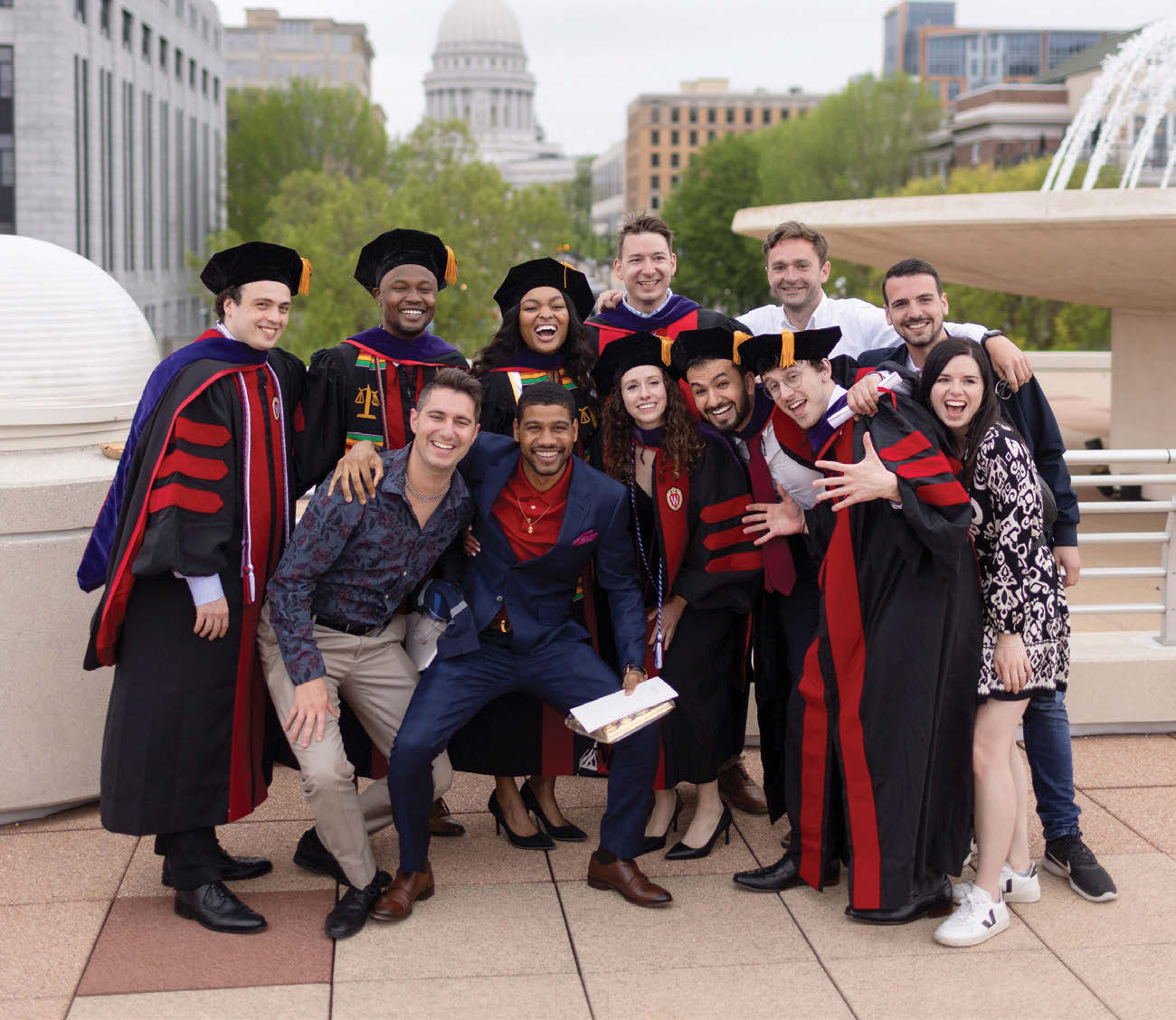 A group of graduates pose together in a photo on the Monona Terrace rooftop with the Madison City Capitol building in the background.