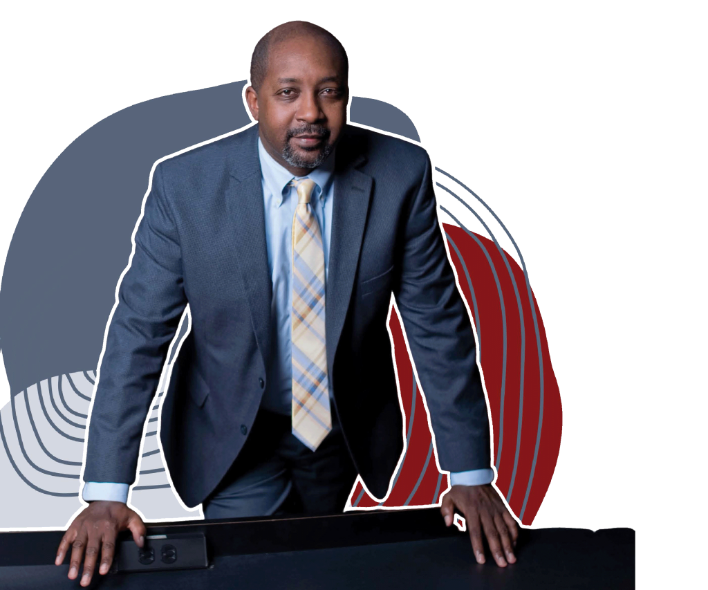A photo of Mario Barnes, a black man wearing a gray blue suit, light blue buttoned shirt, and yellow and blue cross hatched tie, while leaning against a table and has a confident expression.