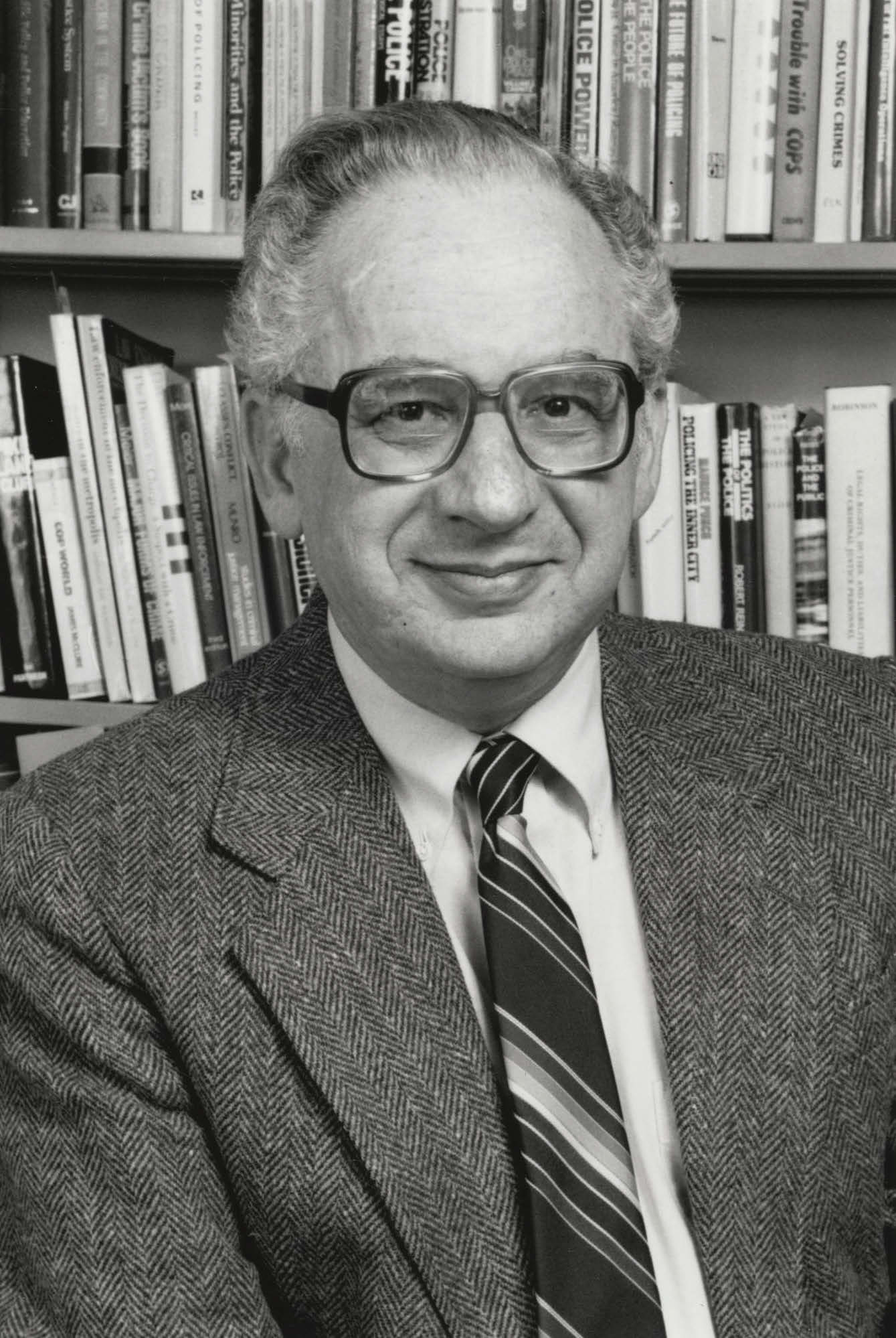 A black and white photo of Herman Goldstein from 1989, provided by the UW Archives. Herman is smiling at the camera while wearing large-framed glasses, a tweed suit jacket, and striped tie.