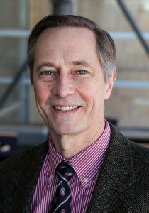 A headshot photo of John Ohnesorge, wearing a twill black suit jacket, with a maroon pinstriped shirt, and UW-themed tie that is mostly black with white stripes and UW logos.