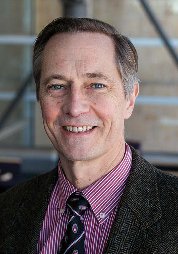 A headshot photo of John Ohnesorge, wearing a twill black suit jacket, with a maroon pinstriped shirt, and UW-themed tie that is mostly black with white stripes and UW logos.