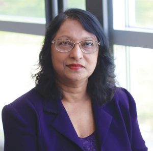 Headshot photo of Sumudu Atapattu, wearing glasses and a purple blazer smiling while looking at the camera in front of windowed space with greenery outside.