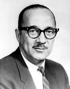A black and white photo of William H. Hastie (1904-1976), wearing black frame glasses, a dark suit jacket, and a striped tie.