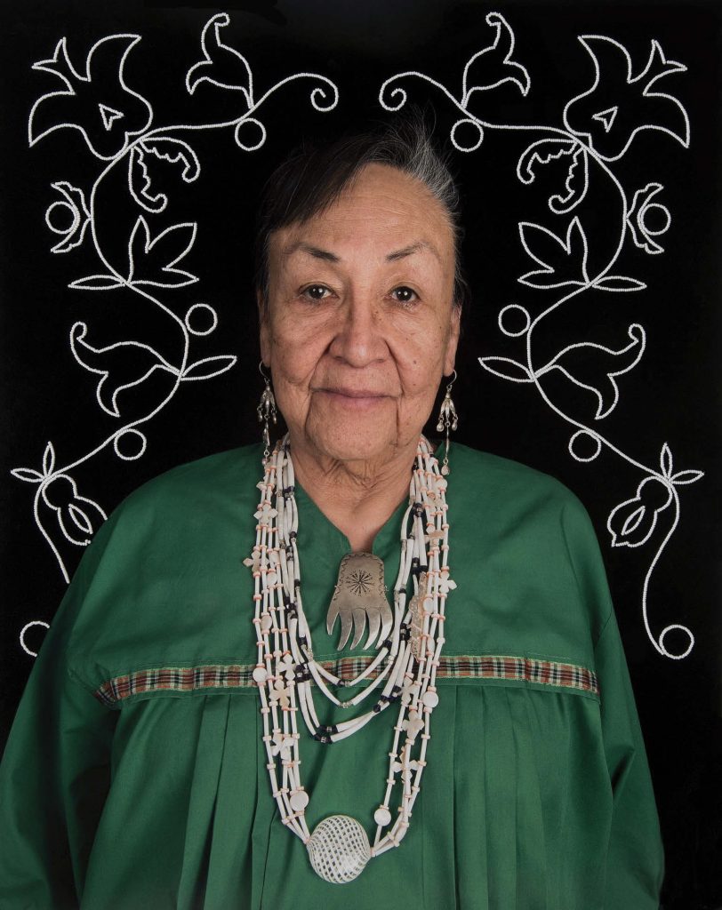 A portrait photo of Judge JoAnn Jones by her son, UW-Madison Professor Tom Jones. She is of native descent and wearing traditional clothing in green and necklaces in white, in front of a black background with white embroidery of flowers.