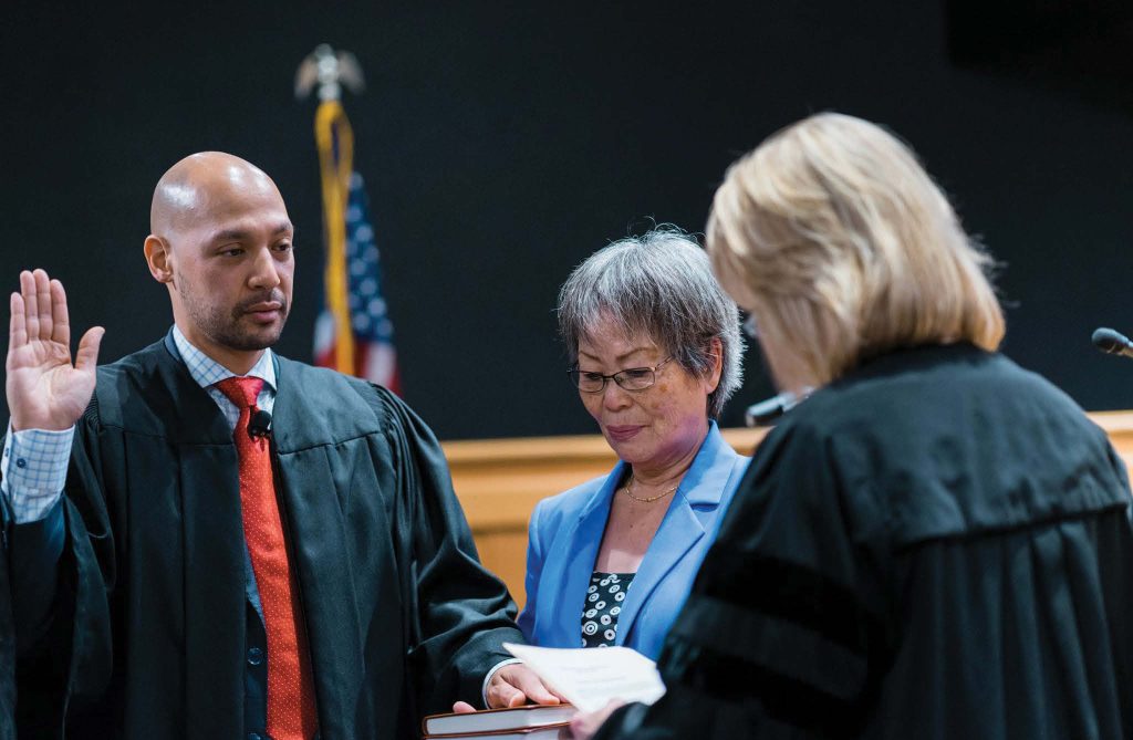 A photo of the swearing in ceremony for Judge Ramon Alvarado, in which he is wearing judge's robes and a red tie, his hand is up with palm facing outward, and he has a pensive expression.