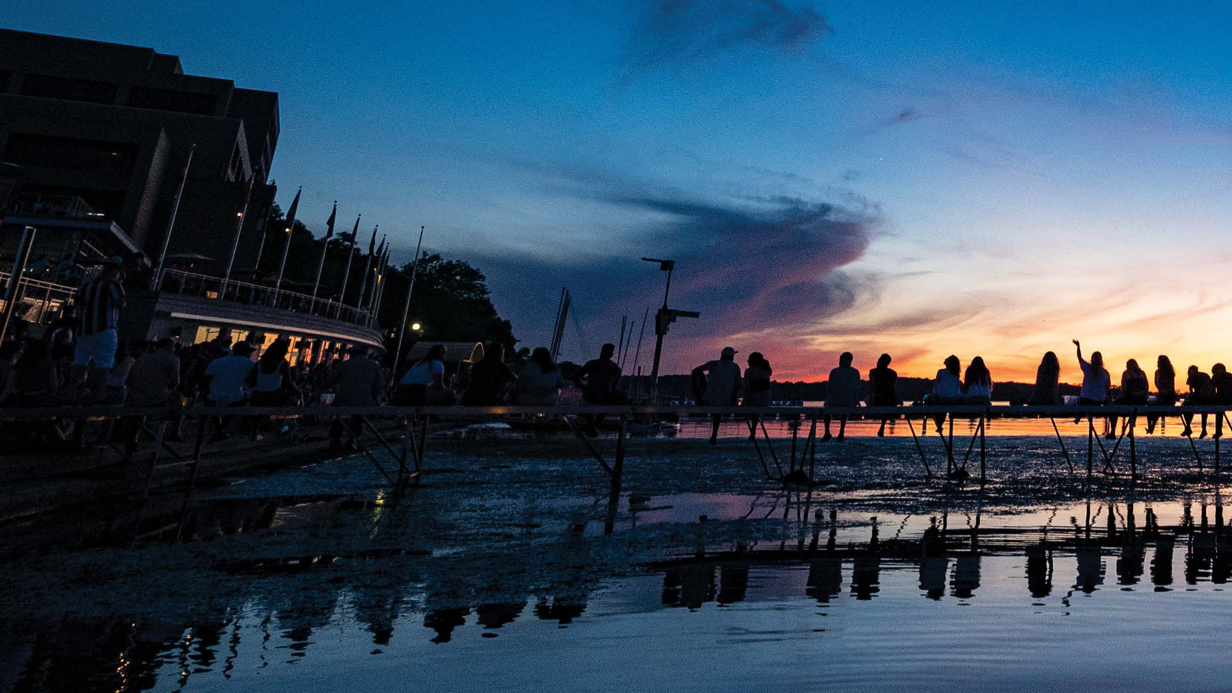 A pleasant photo of the Mondota Terrace in fading light, with docks on which people are seated to view a colorful sunset topped by a deep-blue night sky; the water below reflects it all like a mirror.