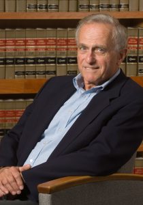 A photo of Frank Tuerkheimer sitting relaxed in a chair while looking at the camera. He sits in front of a large bookshelf of law books.