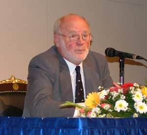 Charles Irish sits at a table on a stage. The table has flowers on top, and he speaks into a microphone. He is bald with white hair and a white beard, wearing glasses.