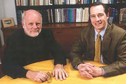 Charles Irish and John Ohnesorge sit at a table in front of a bookshelf. Charles is bald with white hair and white beard and wears a black sweater, while John wears an olive colored suit jacket over a white shirt with yellow tie.