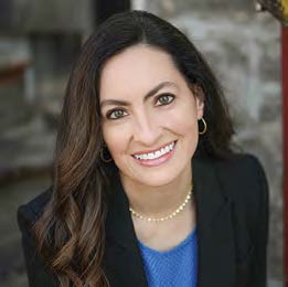 A headshot photo of Rebecca Scheller. She smiles while looking at the camera, wearing a gold necklace, black blazer over a light blue top, against a stone background.