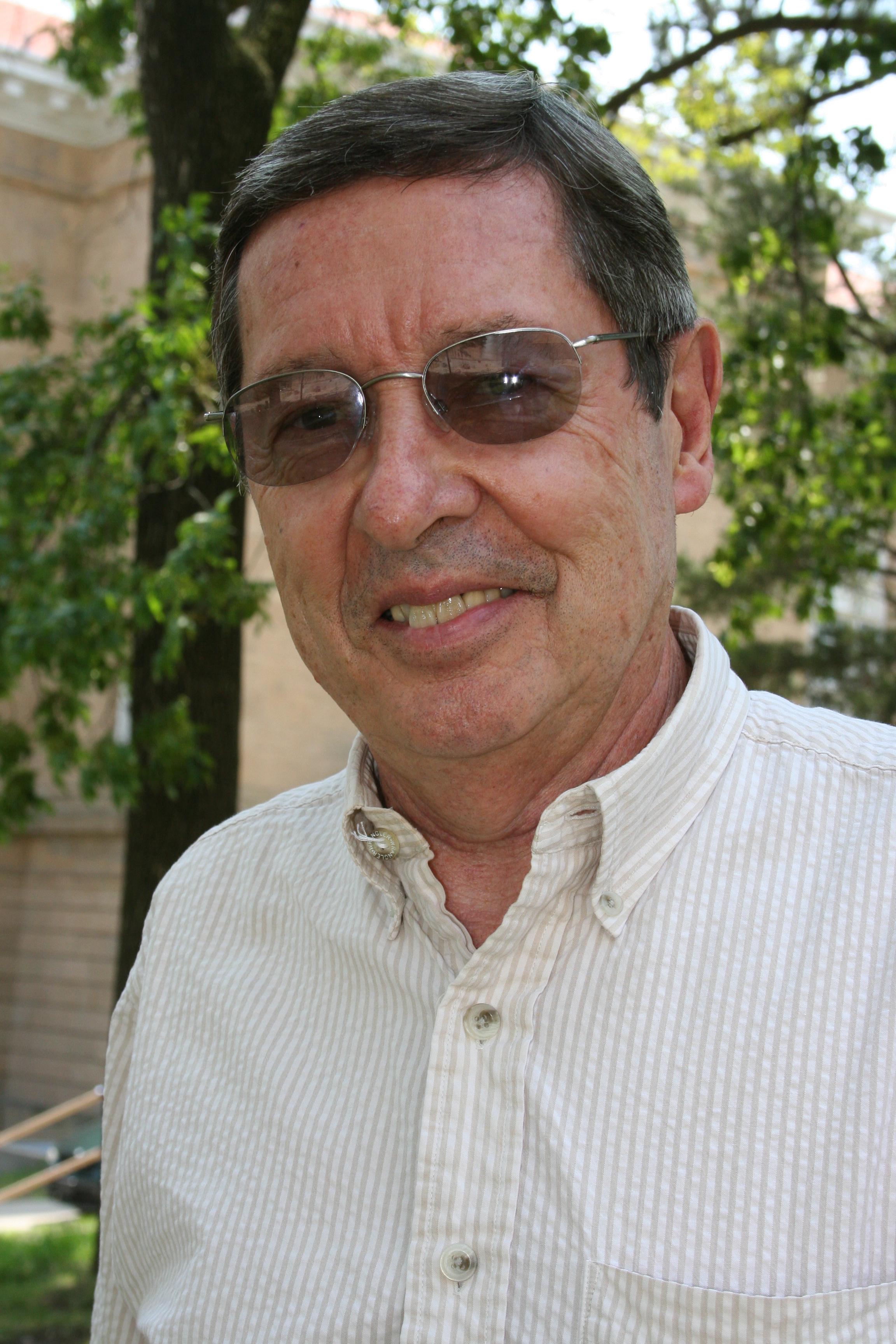 Headshot photo of Joseph Thome wearing sunglasses and a white shirt while smiling at the camera. He is standing in front of a tree with draped leaves.