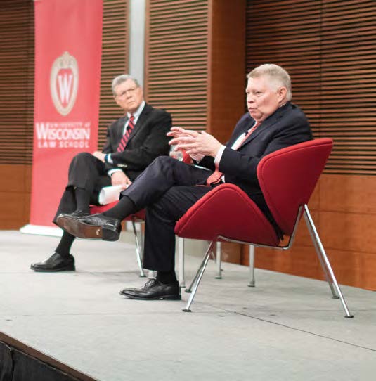 Judge Luttig gestures with open hands while he speaks, seated on stage across from the event moderator.