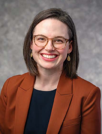 A headshot photo of Renagh O'Leary. She smiles at the camera while wearing tourtoiseshell colored round glasses, teardrop golden earrings, a brown blazer over a dark blue top.