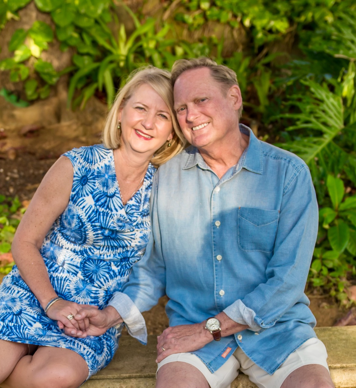 A photo of Joan and Tom Stafslien sitting together in front of green foliage. Joan and Tom rest their heads together with a smile.